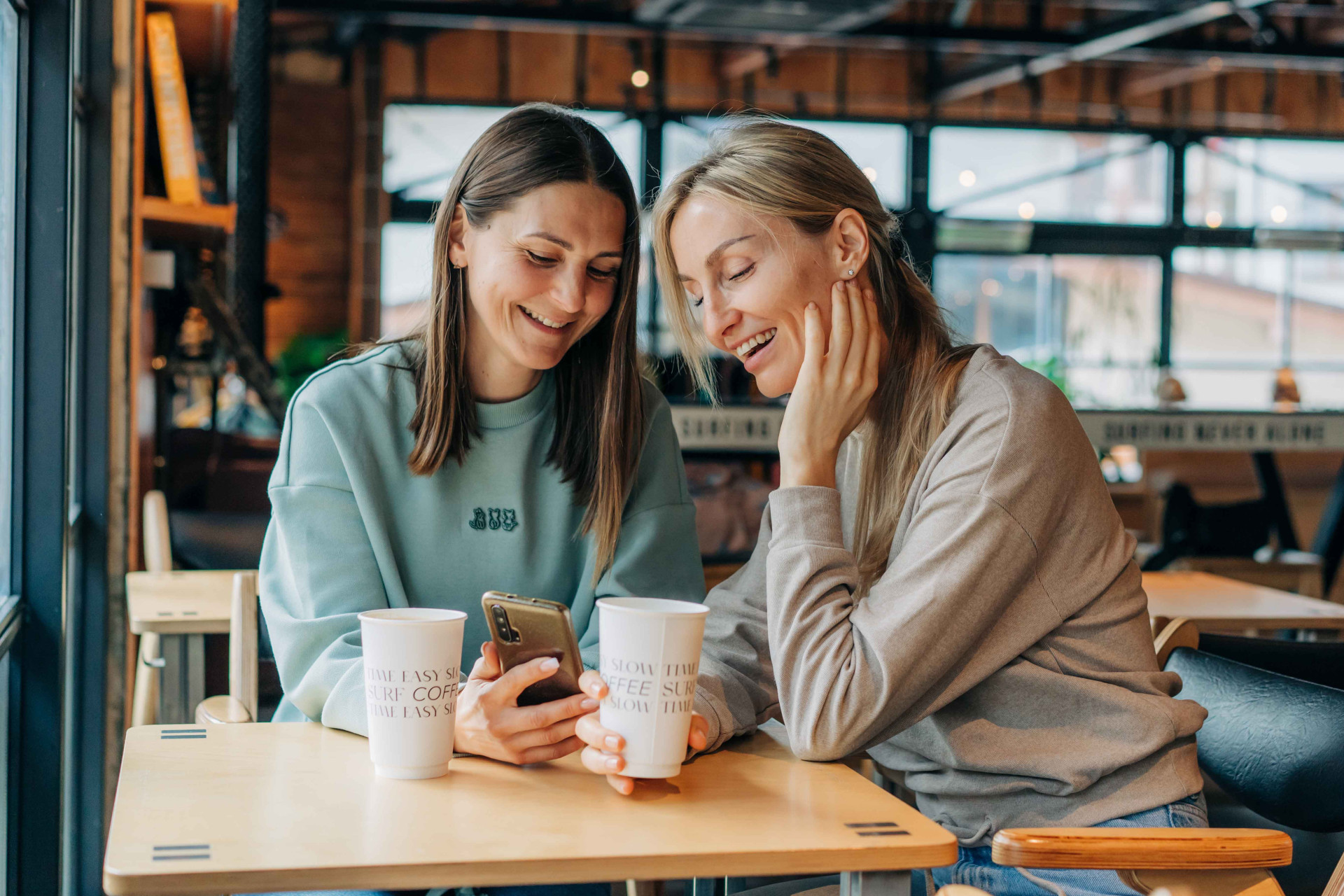 Two women sitting coffee house laughing use phone online communication lr